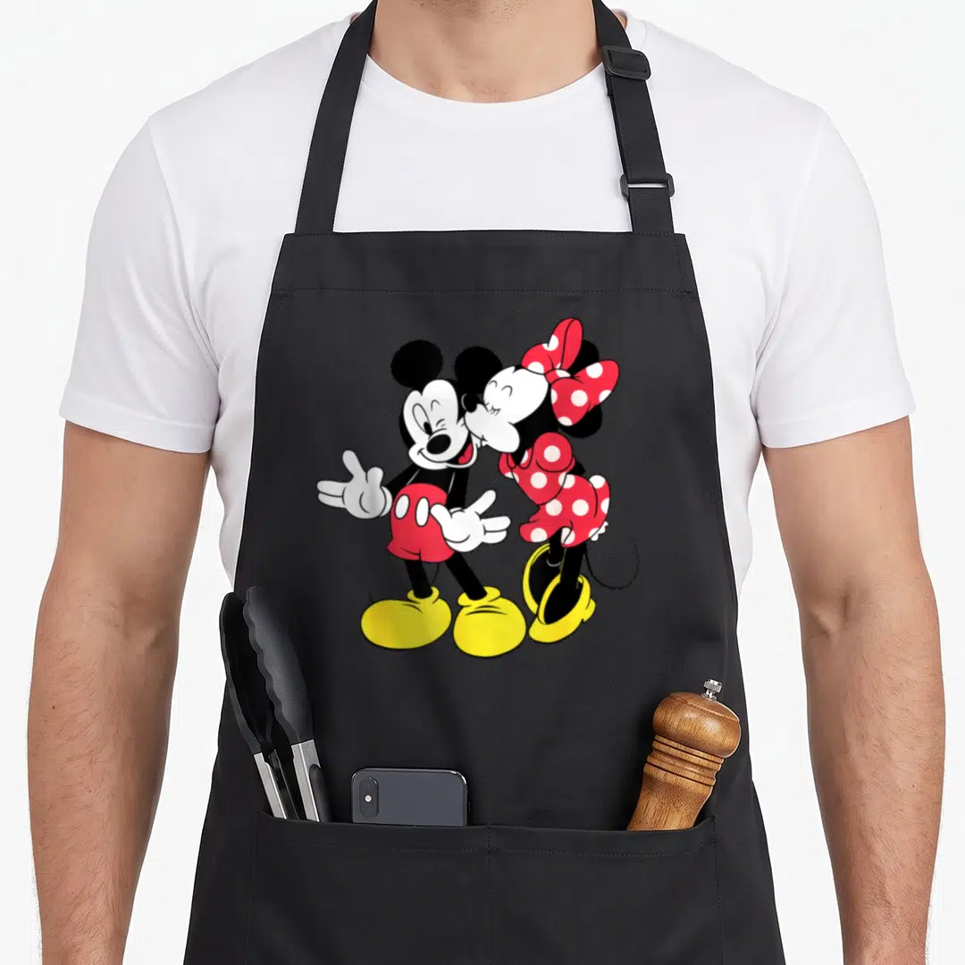Funny mouse lover apron - Mickey and Minnie standing side-by-side cartoon kiss in black red yellow white — shown on a black cotton canvas apron against a white background