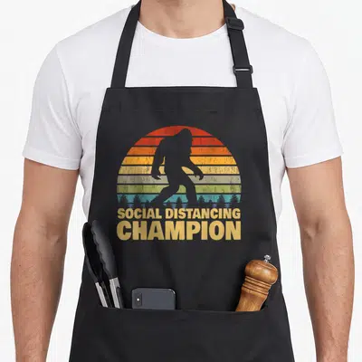 Social trendy apron - Grey bigfoot silhouette over multi-colored striped semi-circle with stacked yellow text reading — shown on a black cotton canvas apron against a white background
