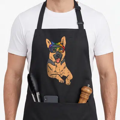 Shepherd bandana gay pride apron - Cartoon tan and black dog wearing rainbow glasses and bandana — shown on a black cotton canvas apron against a white background