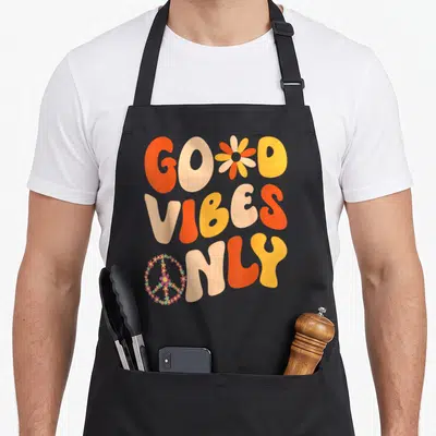 Peace 60s tie-dye apron - Retro text Good Vibes Only with flower and peace sign graphic in orange and peach — shown on a black cotton canvas apron against a white background