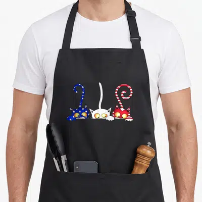 Cat flag 4th of july apron - Three cartoon cats in blue white and red peeking up with starry and striped tails — shown on a black cotton canvas apron against a white background