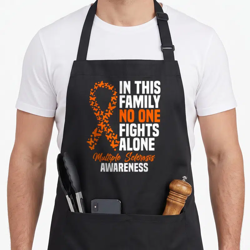 Multiple sclerosis ribbon apron - Orange butterfly awareness ribbon next to stacked white text reading In This Family — shown on a black cotton canvas apron against a white background