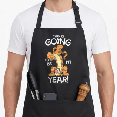 Tigger friends apron - Upside down orange cartoon tiger with white text THIS IS GOING TO BE MY YEAR! — shown on a black cotton canvas apron against a white background