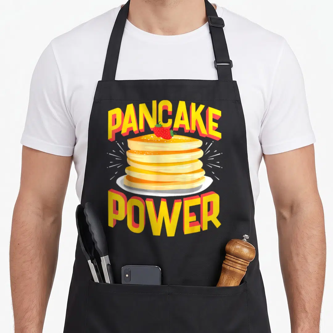 Power pancake apron - Cartoon stack of pancakes with red strawberries and arched yellow text reading PANCAKE POWER — shown on a black cotton canvas apron against a white background
