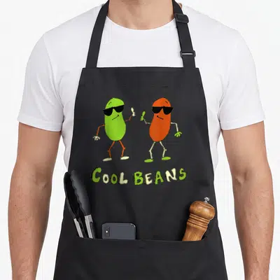 Bean 80s apron - Two cartoon beans with sunglasses above COOL BEANS text in green and yellow — shown on a black cotton canvas apron against a white background