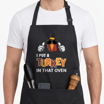 Pregnancy turkey thanksgiving apron - Cartoon turkey head with sunglasses and thumbs up above stacked text I PUT — shown on a black cotton canvas apron against a white background