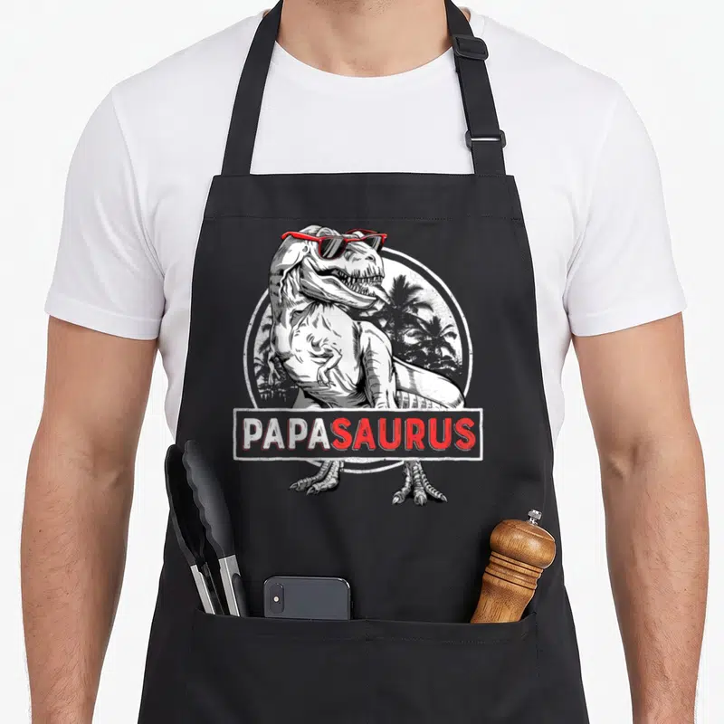 Rex papa apron - T-Rex with red sunglasses and PAPASAURUS text over palm trees — shown on a black cotton canvas apron against a white background