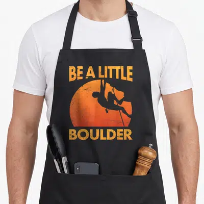 Pun boulder apron - Distressed orange text BE A LITTLE BOULDER surrounding a climber silhouette inside a sun circle — shown on a black cotton canvas apron against a white background