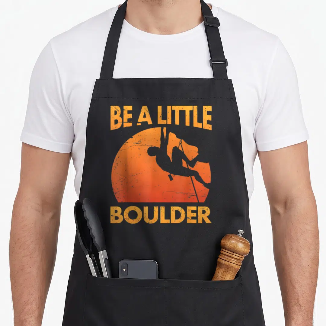 Pun boulder apron - Distressed orange text BE A LITTLE BOULDER surrounding a climber silhouette inside a sun circle — shown on a black cotton canvas apron against a white background