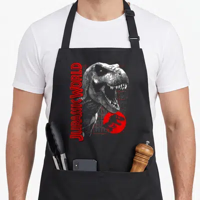 Grid t-rex apron - Vertical JURASSICWORLD text beside a roaring T-Rex head and small circular dinosaur logo — shown on a black cotton canvas apron against a white background