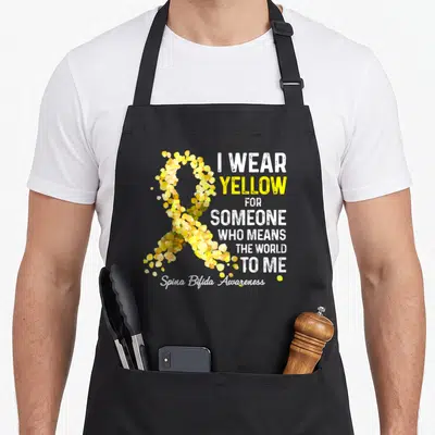 Someone special aprons - Yellow confetti ribbon and text I WEAR YELLOW FOR SOMEONE WHO MEANS THE WORLD TO ME — shown on a black cotton canvas apron against a white background
