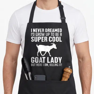Pygmy goat apron - White goat silhouette between I never dreamed super cool goat lady killing it text — shown on a black cotton canvas apron against a white background