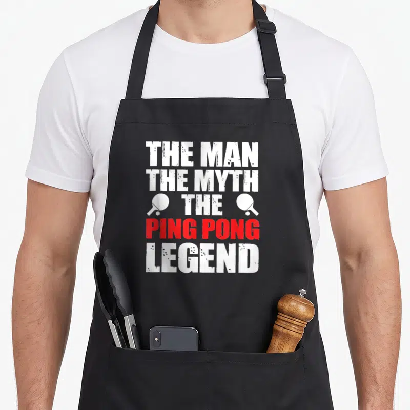 Myth pong apron - Stacked text reading THE MAN THE MYTH THE PING PONG LEGEND flanked by two paddles — shown on a black cotton canvas apron against a white background