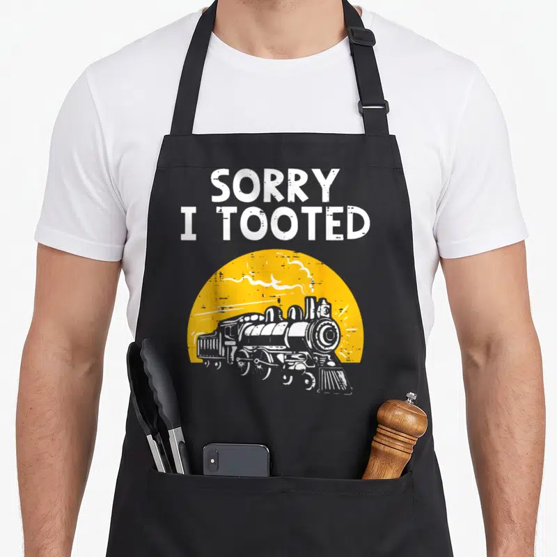 Sorry boy train apron - White steam locomotive in front of yellow semi-circle with SORRY I TOOTED text — shown on a black cotton canvas apron against a white background