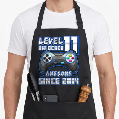 11th birthday level gamer video game apron - LEVEL 11 UNLOCKED AWESOME SINCE 2014 text surrounding a blue and grey game — shown on a black cotton canvas apron against a white background
