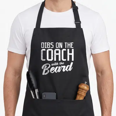 Football beard apron - White stacked text reading DIBS ON THE COACH with the Beard on grey background — shown on a black cotton canvas apron against a white background
