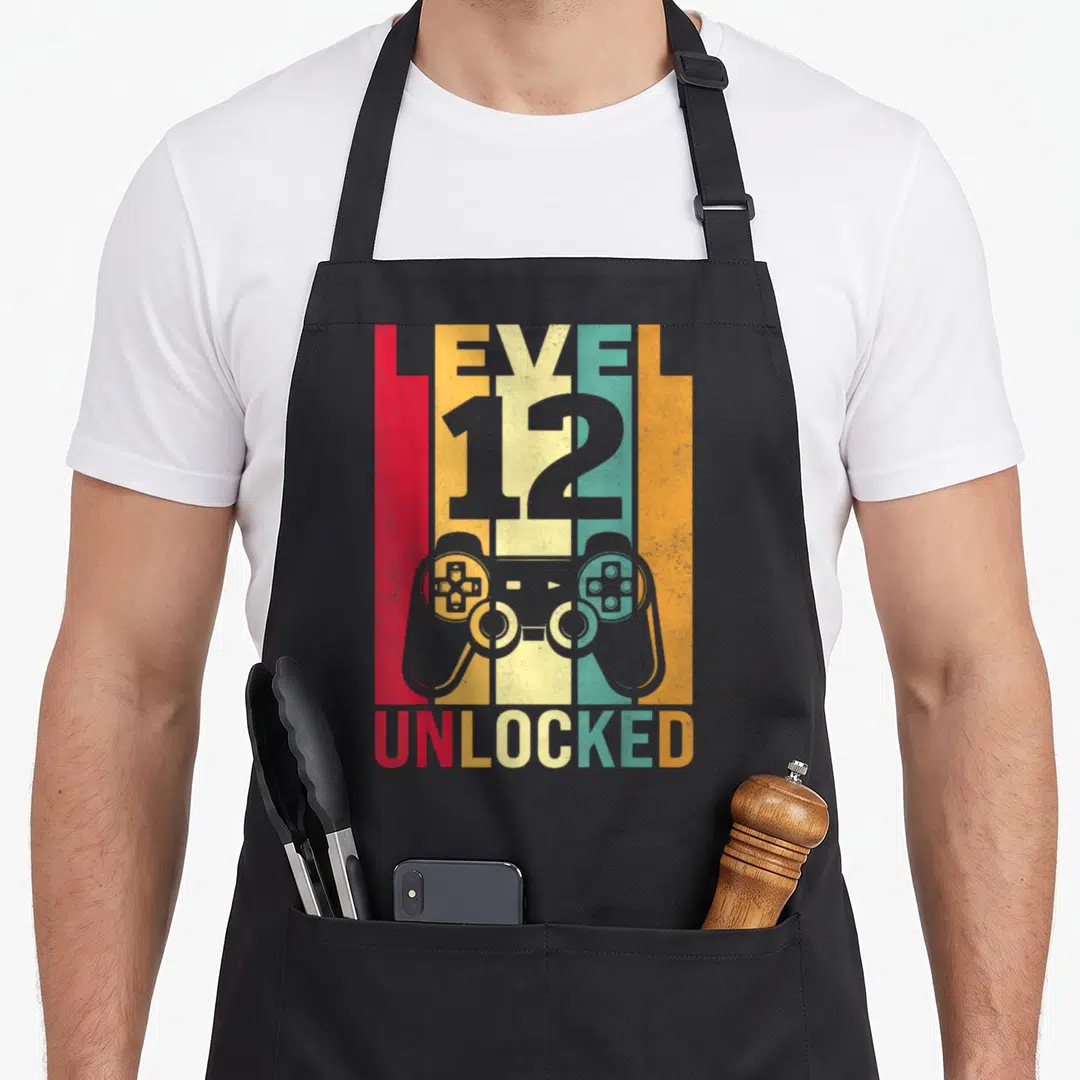 12yr dad and son aprons - Retro game controller over five red yellow cream teal orange stripes with LEVEL 12 UNLOCKED — shown on a black cotton canvas apron against a white background