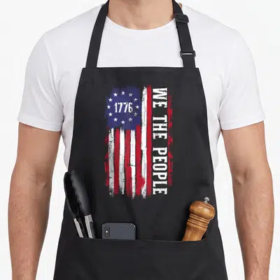 Independence american flag apron - Distressed USA flag with 1776 in blue canton and vertical WE THE PEOPLE text — shown on a black cotton canvas apron against a white background