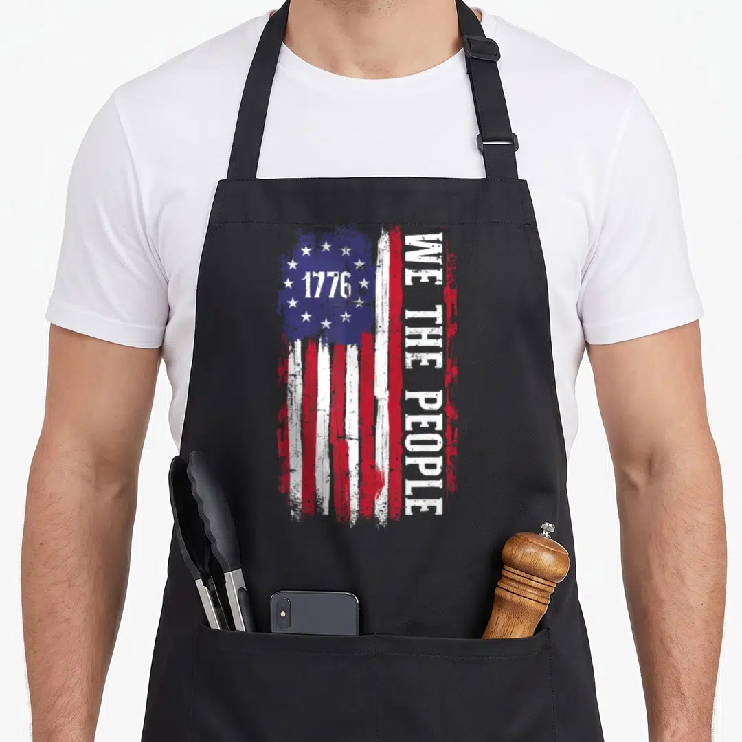 Independence american flag apron - Distressed USA flag with 1776 in blue canton and vertical WE THE PEOPLE text — shown on a black cotton canvas apron against a white background