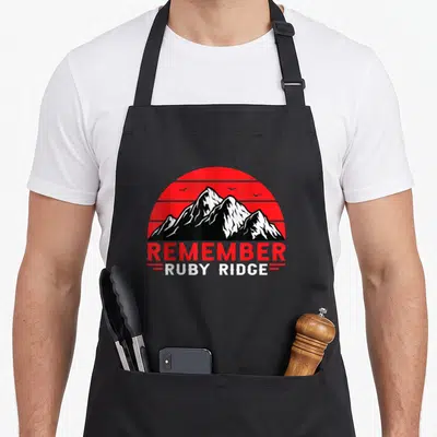 Remember the ruby apron - Red striped sun and black white mountains with REMEMBER RUBY RIDGE text — shown on a black cotton canvas apron against a white background