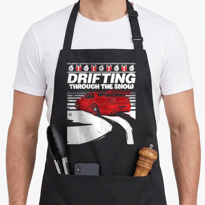 Car design imports vintage apron - Red car drifting left with DRIFTING THROUGH THE SNOW text and turbo gift box row — shown on a black cotton canvas apron against a white background