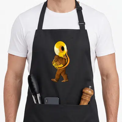 Sousaphone bigfoot apron - Cartoon brown bigfoot standing playing a large yellow sousaphone instrument — shown on a black cotton canvas apron against a white background