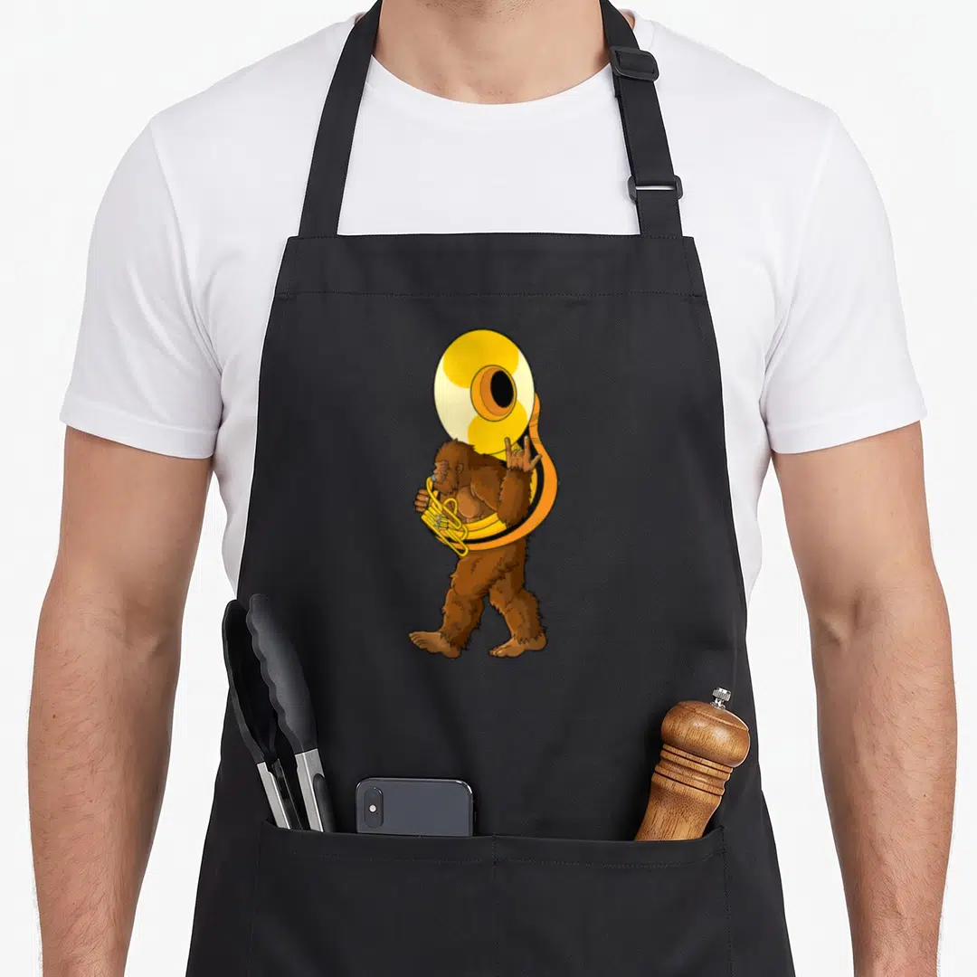 Sousaphone bigfoot apron - Cartoon brown bigfoot standing playing a large yellow sousaphone instrument — shown on a black cotton canvas apron against a white background