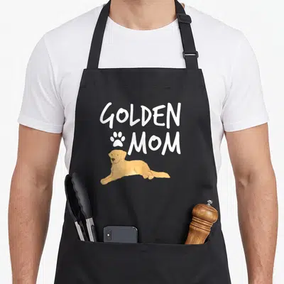 Dog puppy golden retriever apron - Cartoon golden dog lying under white GOLDEN MOM text and paw print — shown on a black cotton canvas apron against a white background
