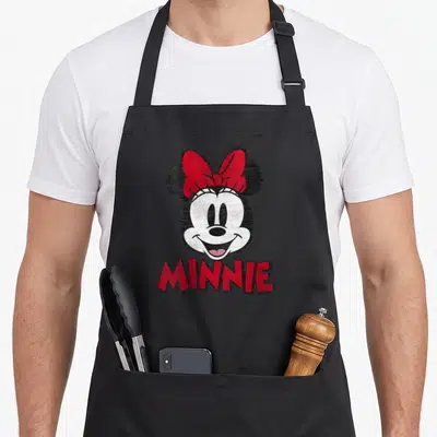 Portrait friends apron - Minnie Mouse head above red MINNIE text in black white and red cartoon style — shown on a black cotton canvas apron against a white background