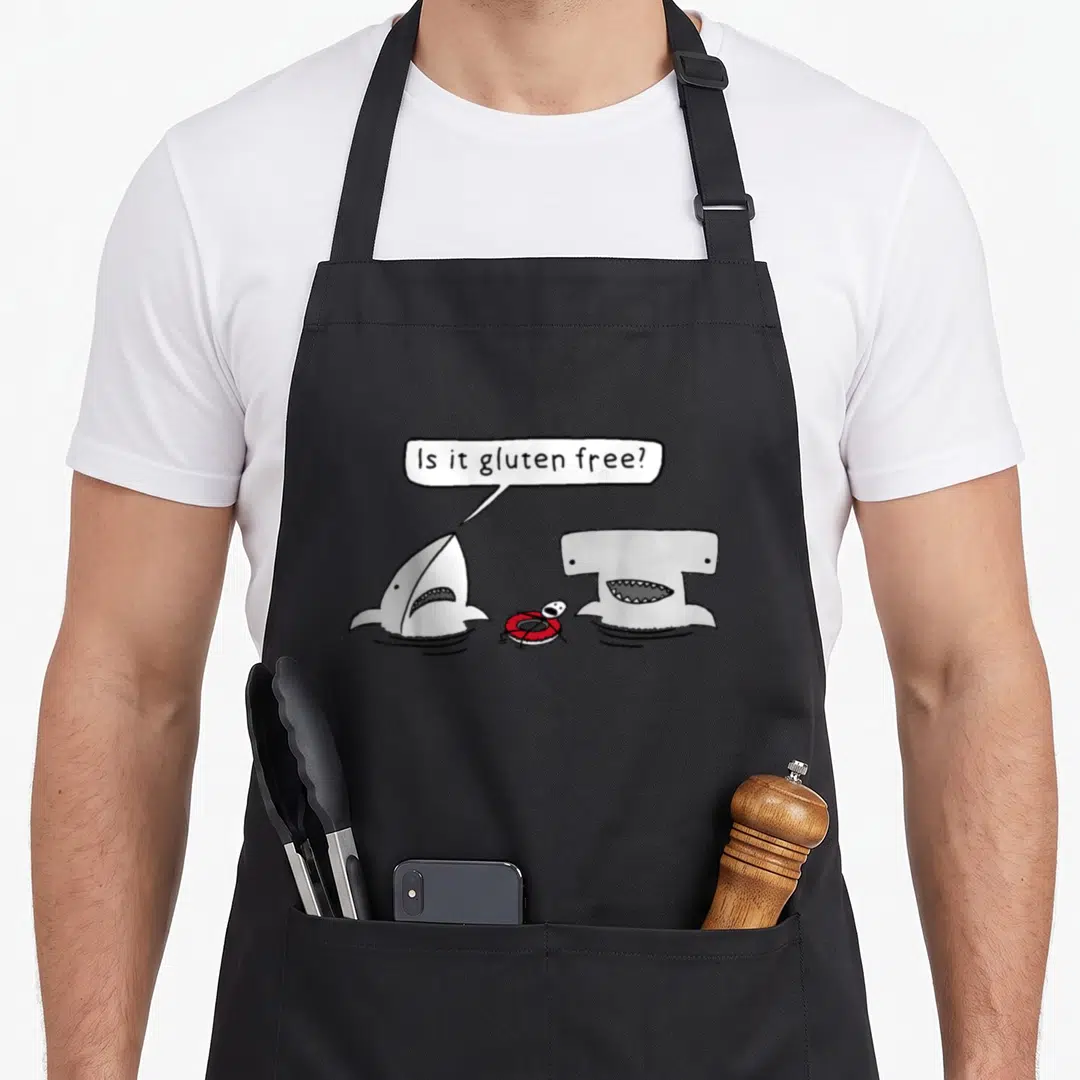 Gluten graphical apron - Two sharks face swimmer in lifebuoy, speech bubble reads Is it gluten free — shown on a black cotton canvas apron against a white background