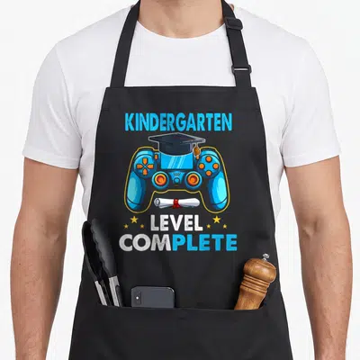 School kindergarten apron - Blue game controller with graduation cap, diploma, and KINDERGARTEN LEVEL COMPLETE text — shown on a black cotton canvas apron against a white background