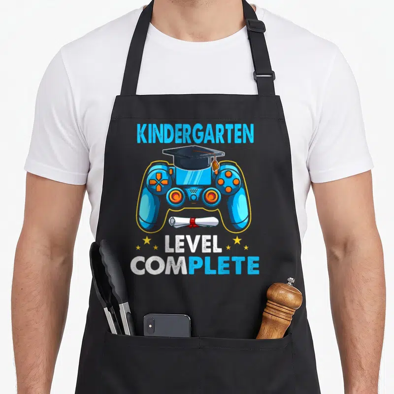 School kindergarten apron - Blue game controller with graduation cap, diploma, and KINDERGARTEN LEVEL COMPLETE text — shown on a black cotton canvas apron against a white background