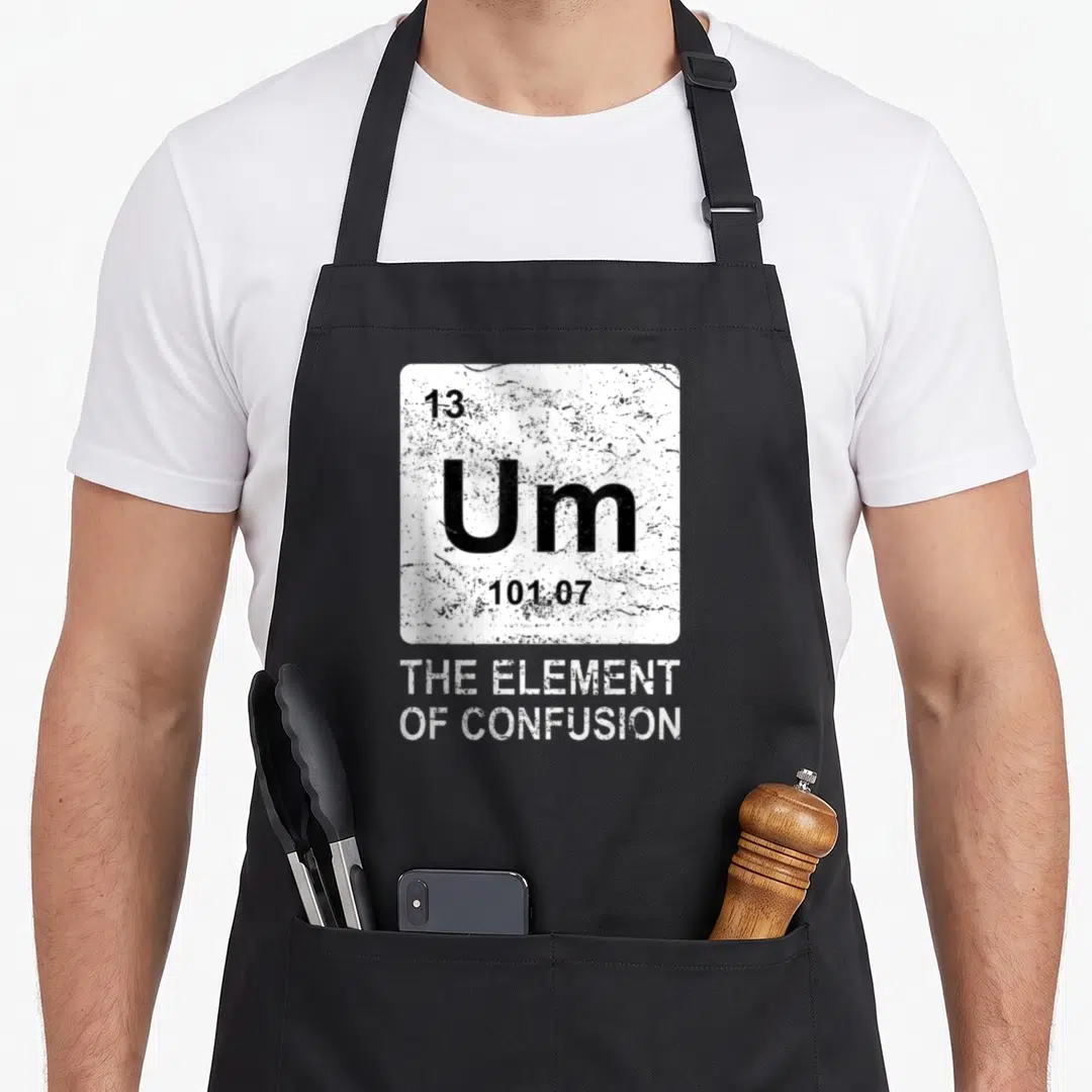 Confusion chemistry apron - White tile with Um 13 101.07 and THE ELEMENT OF CONFUSION in distressed white text on black — shown on a black cotton canvas apron against a white background