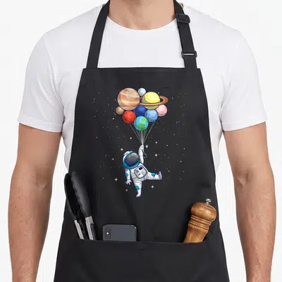 Space themed aprons - Cartoon astronaut holding planet balloons in white blue yellow red stars — shown on a black cotton canvas apron against a white background