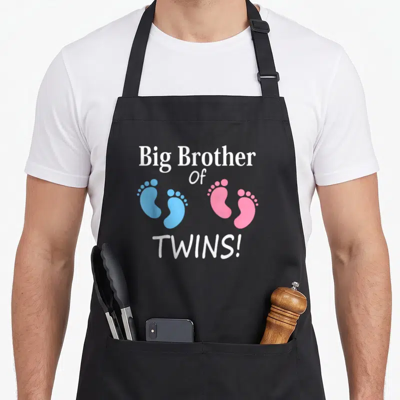 Brother twins apron - White Big Brother Of TWINS text above two blue and two pink baby footprints — shown on a black cotton canvas apron against a white background