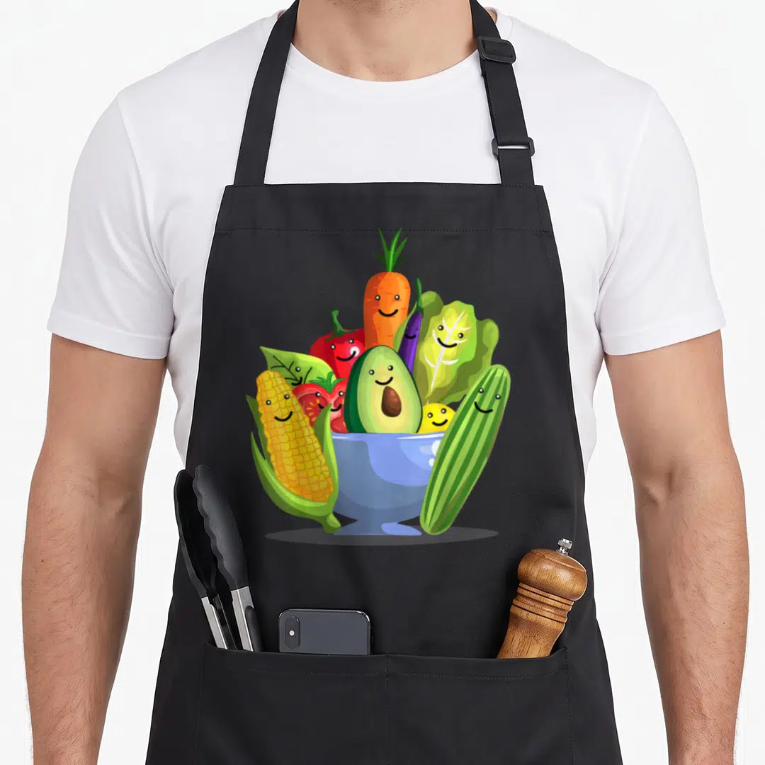 Vegetarian vegan veggie apron - Cartoon smiling vegetables including carrot corn and avocado in a blue bowl — shown on a black cotton canvas apron against a white background
