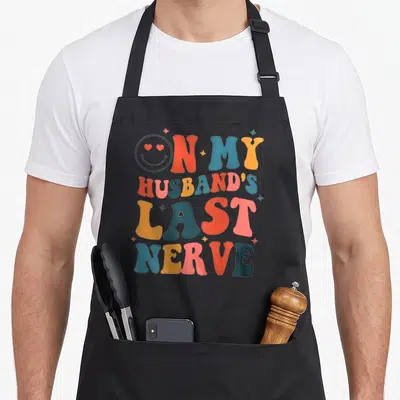 Nerve husband apron - Retro text design ON MY HUSBAND'S LAST NERVE in red blue yellow pink with smiley face — shown on a black cotton canvas apron against a white background