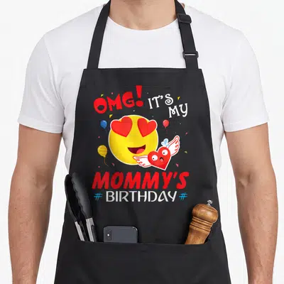 Smile mother's apron - Heart eyes emoji holding winged heart with balloons and OMG It's My Mommy's Birthday text — shown on a black cotton canvas apron against a white background