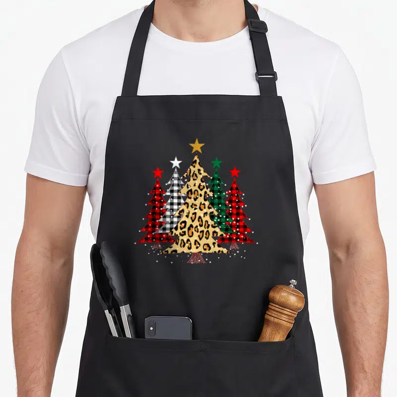Buffalo merry christmas apron - Five Christmas trees with leopard, plaid, and star toppers and white snow dots — shown on a black cotton canvas apron against a white background