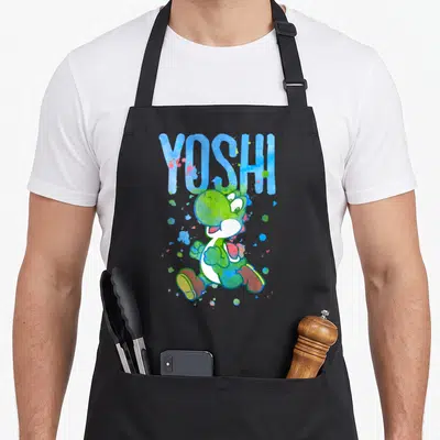 Watercolor splash aprons - YOSHI text above green cartoon dinosaur with blue, green, white, and red paint splatters — shown on a black cotton canvas apron against a white background