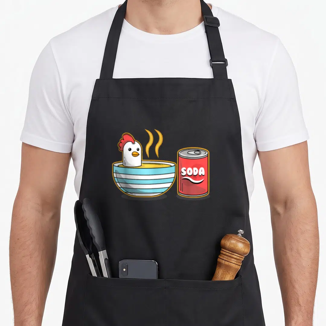 Soup chicken apron - Cartoon chicken head in striped bowl next to red soda can with SODA text — shown on a black cotton canvas apron against a white background