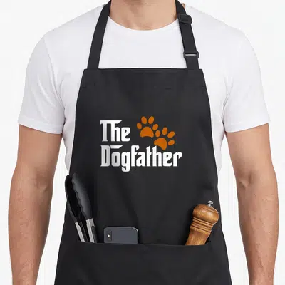 Father dog dad apron - The Dogfather text with two overlapping orange paw prints — shown on a black cotton canvas apron against a white background
