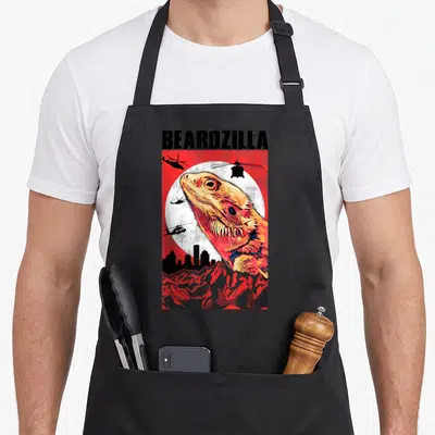 Retro bearded dragon apron - Beardzilla text above bearded dragon head, helicopters, and city skyline on red background — shown on a black cotton canvas apron against a white background