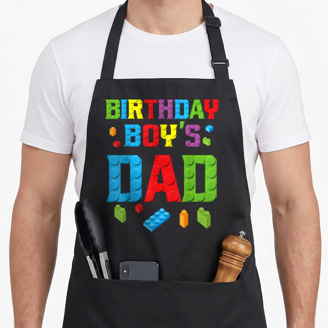 Builder birthday block & apron - Colorful BIRTHDAY BOY'S DAD text made of toy bricks with scattered pieces — shown on a black cotton canvas apron against a white background