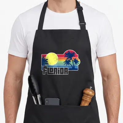 Sunshine florida apron - Retro Florida sunset with yellow sun, striped background, blue palms, and white text — shown on a black cotton canvas apron against a white background