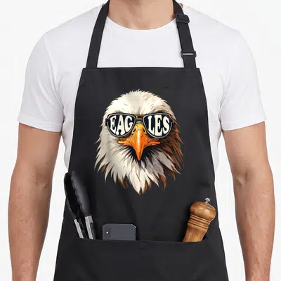 Sunglass eagles grill apron - Realistic eagle head wearing sunglasses with EAGLES text in lenses, black bib apron — shown on a black cotton canvas apron against a white background