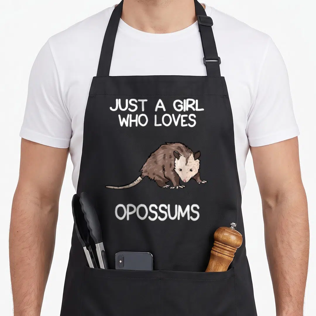 Owner opossum apron - Cartoon brown and grey opossum between white text Just a Girl Who Loves Opossums — shown on a black cotton canvas apron against a white background