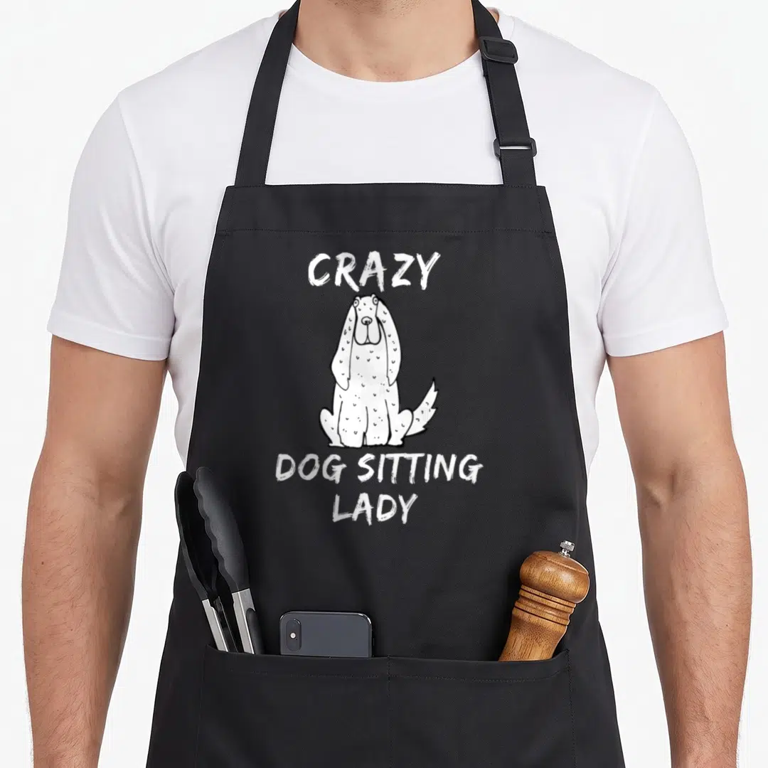 Sitting dog cooking apron - White cartoon dog centered between arched CRAZY text and stacked DOG SITTING LADY lettering — shown on a black cotton canvas apron against a white background
