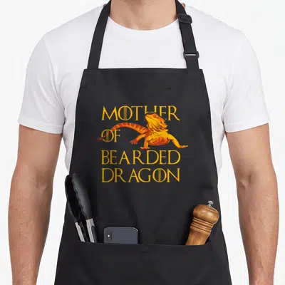 Bearded mother's apron - Cartoon bearded dragon with stacked yellow text reading Mother of Bearded Dragon — shown on a black cotton canvas apron against a white background