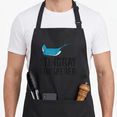 Stingray whisperer funny apron - Blue stingray illustration above stacked black text reading STINGRAY WHISPERER on grey — shown on a black cotton canvas apron against a white background